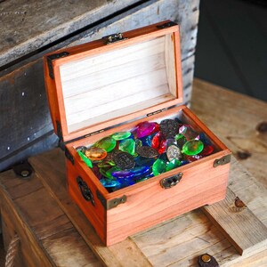 May include: An open wooden treasure chest filled with colorful glass gems and dark metal coins. The chest is made of reddish-brown wood with metal corner brackets and a clasp. The gems are various colors, including red, green, blue, and yellow.