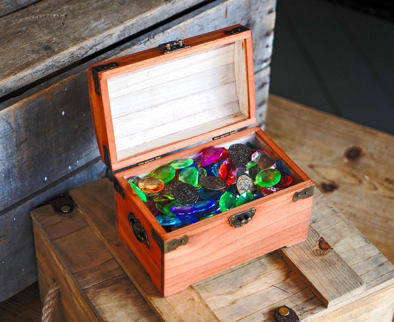 May include: An open wooden treasure chest filled with colorful glass gems and dark metal coins. The chest is made of reddish-brown wood with metal corner brackets and a clasp. The gems are various colors, including red, green, blue, and yellow.