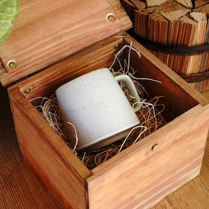 May include: A speckled off-white ceramic mug with a handle rests inside a square wooden box filled with thin wood shavings. The box features a hinged lid with metal hardware and a natural wood grain finish, presenting a clean and simple aesthetic.