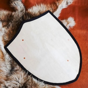 May include: A heater-shaped wooden shield with a light-coloured, smooth surface and a thick black border. The shield features four small, evenly spaced circular metal rivets or fasteners embedded into the wood, arranged in a rectangular pattern.