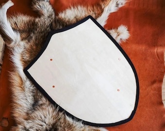 May include: A heater-shaped wooden shield with a light-coloured, smooth surface and a thick black border. The shield features four small, evenly spaced circular metal rivets or fasteners embedded into the wood, arranged in a rectangular pattern.