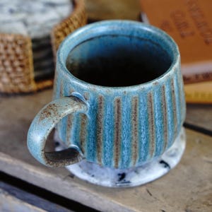 May include: A ceramic mug with a blue and brown reactive glaze finish featuring vertical ribbed textures along the exterior body. The mug has a curved handle and a wide circular opening, showcasing a rustic aesthetic with visible colour variations.