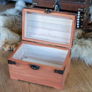 May include: Open wooden treasure chest with a reddish-brown finish, black metal hinges, and a clasp. The interior is lined with light-colored wood. Another chest is visible in the background. The chest is on a wooden surface.