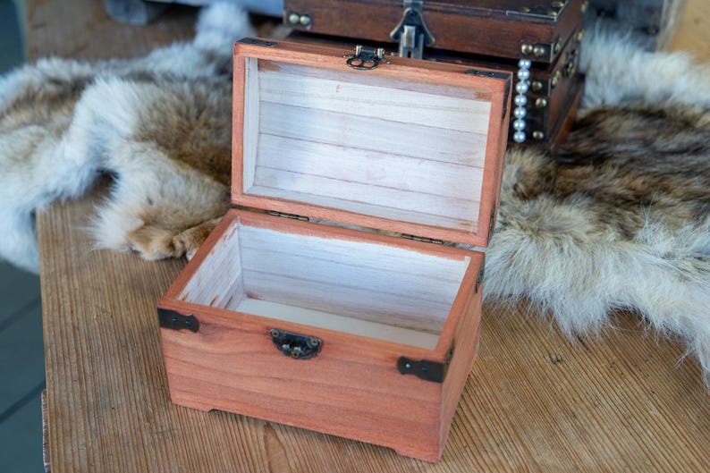 May include: Open wooden treasure chest with a reddish-brown finish, black metal hinges, and a clasp. The interior is lined with light-colored wood. Another chest is visible in the background. The chest is on a wooden surface.
