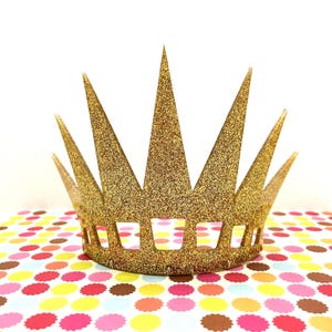 May include: A gold glitter crown featuring a series of tall, pointed spikes extending upward from a solid base. The base of the crown is decorated with a row of rectangular cutouts, and the entire surface is covered in a dense, sparkling gold texture.