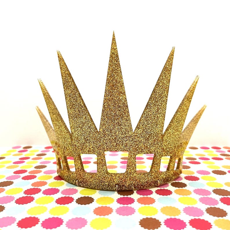 May include: A gold glitter crown featuring a series of tall, pointed spikes extending upward from a solid base. The base of the crown is decorated with a row of rectangular cutouts, and the entire surface is covered in a dense, sparkling gold texture.