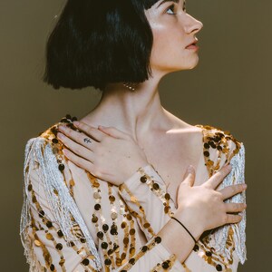 May include: A beige long-sleeved top adorned with vertical rows of shimmering gold sequins and silver fringe detailing on the shoulders, paired with a silver glitter crown featuring pointed spikes worn on dark hair.