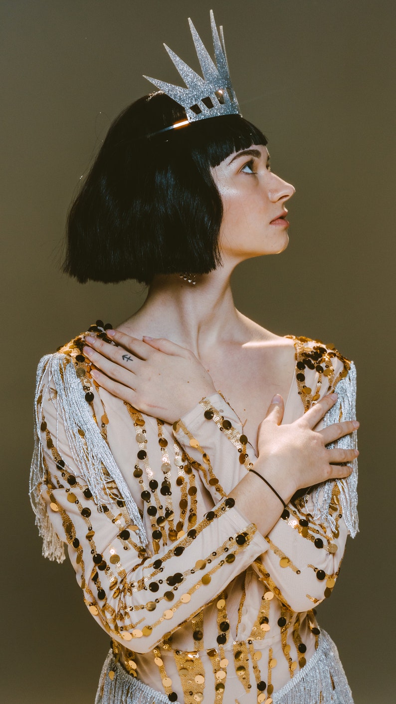 May include: A beige long-sleeved top adorned with vertical rows of shimmering gold sequins and silver fringe detailing on the shoulders, paired with a silver glitter crown featuring pointed spikes worn on dark hair.