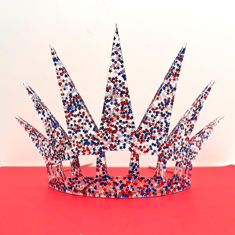 May include: A clear, pointed crown featuring a dense pattern of embedded red, white, and blue star-shaped glitter. The headpiece has a circular base with rectangular cutouts and seven tall, triangular spikes extending upward from the band.