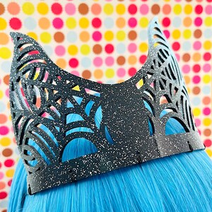 May include: A black, decorative headpiece with a speckled finish and intricate cut-out designs. The headpiece is worn over bright blue hair, with a colorful polka dot background.