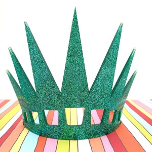 May include: A green glittery crown featuring seven tall, pointed spikes of varying heights and a series of rectangular cutouts along the base. The surface is covered in a dense, sparkling texture that reflects light across the entire structure.
