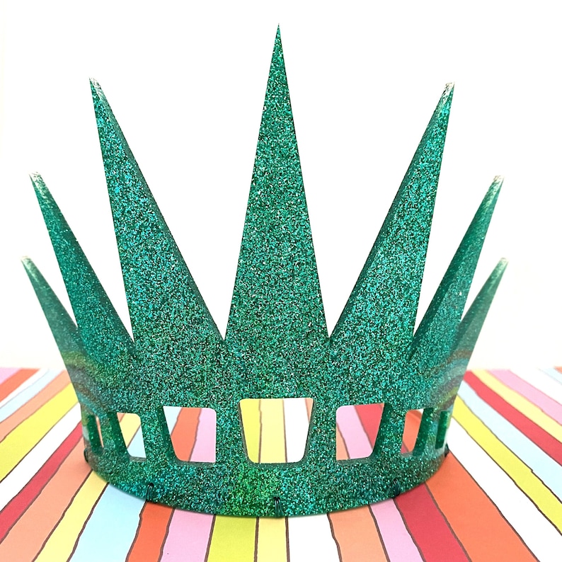 May include: A green glittery crown featuring seven tall, pointed spikes of varying heights and a series of rectangular cutouts along the base. The surface is covered in a dense, sparkling texture that reflects light across the entire structure.