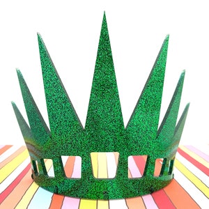 May include: A bright green glitter crown featuring seven tall, pointed spikes and a series of rectangular cutouts along the base. The crown has a textured, sparkling surface that reflects light, creating a vibrant appearance across the entire structure.