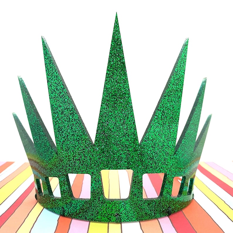 May include: A bright green glitter crown featuring seven tall, pointed spikes and a series of rectangular cutouts along the base. The crown has a textured, sparkling surface that reflects light, creating a vibrant appearance across the entire structure.