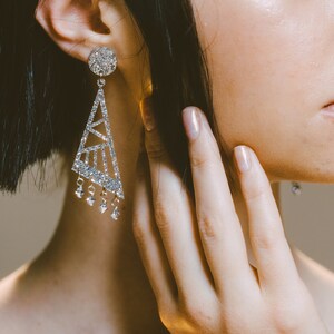 Puede incluir: Un brillante pendiente geométrico con un perno redondo y una caída triangular. La caída presenta un diseño recortado y cristales colgantes. El pendiente es plateado y capta la luz, creando un efecto glamuroso. La imagen muestra un primer plano del pendiente.