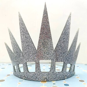 May include: A silver glitter crown featuring a series of tall, pointed triangular spikes extending upward from a solid base. The lower section of the crown includes a row of evenly spaced rectangular cutouts, creating a structured, geometric design.