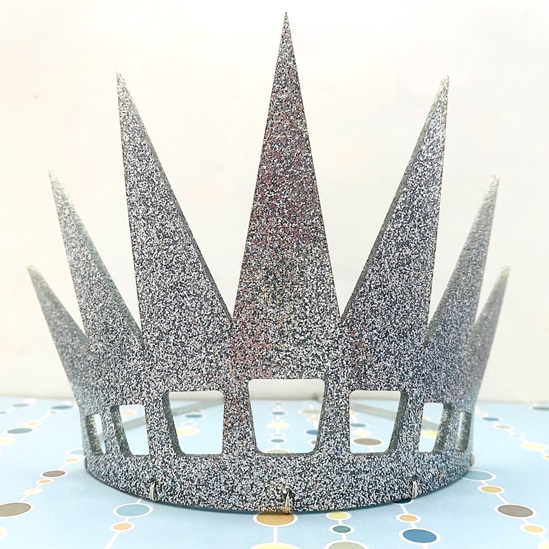 May include: A silver glitter crown featuring a series of tall, pointed triangular spikes extending upward from a solid base. The lower section of the crown includes a row of evenly spaced rectangular cutouts, creating a structured, geometric design.