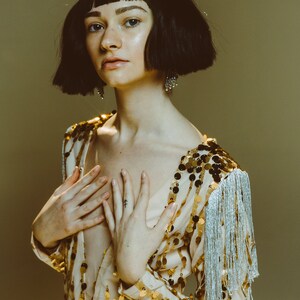 May include: A beige bodysuit featuring vertical rows of gold sequins and silver fringe trim on the sleeves and waist. The garment is paired with a silver glittered crown with tall, pointed spikes, worn over a short, dark, blunt-cut bob hairstyle.
