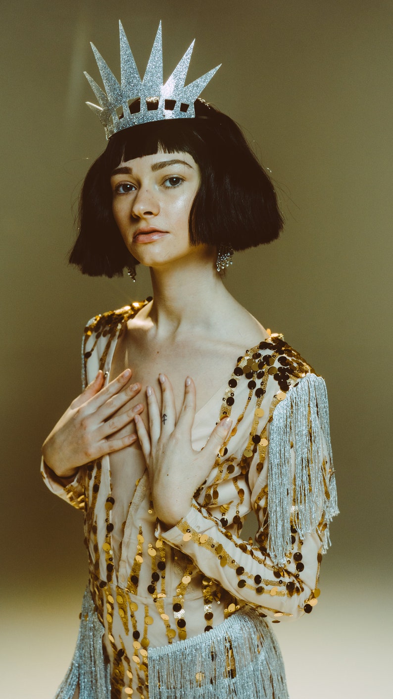 May include: A beige bodysuit featuring vertical rows of gold sequins and silver fringe trim on the sleeves and waist. The garment is paired with a silver glittered crown with tall, pointed spikes, worn over a short, dark, blunt-cut bob hairstyle.