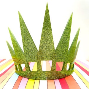 May include: A bright green glittery crown featuring seven tall, pointed spikes and a series of rectangular cutouts along the base. The crown has a textured, sparkling finish that reflects light across its surface, creating a vibrant appearance.