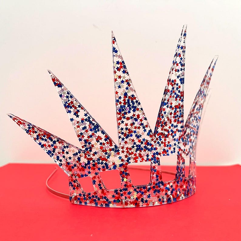 May include: A clear acrylic crown headband featuring a pointed, multi-spiked design. The entire surface is embedded with a dense pattern of small, reflective red, white, and blue star-shaped glitter, creating a vibrant, patriotic, and sparkling effect.
