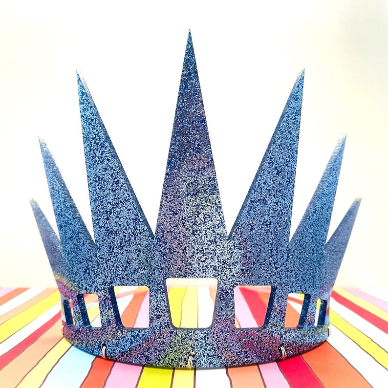 May include: A blue crown with a glittery finish and a series of tall, pointed spikes. The base of the crown features a row of rectangular cutouts, and the entire surface is covered in a dense, sparkling blue texture that reflects light.