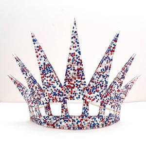 May include: A crown featuring seven pointed spikes and a series of rectangular cutouts along the base. The entire structure is made of a clear material embedded with a dense pattern of small red, white, and blue star-shaped glitter.
