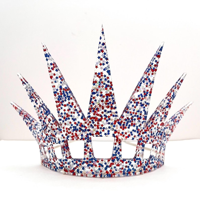 May include: A crown featuring seven pointed spikes and a series of rectangular cutouts along the base. The entire structure is made of a clear material embedded with a dense pattern of small red, white, and blue star-shaped glitter.
