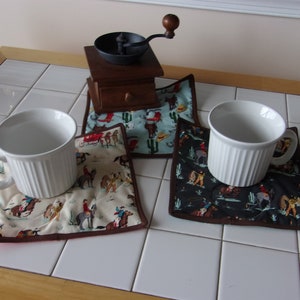 May include: Three white mugs rest on patterned quilted coasters with a brown border. The coasters feature cowboy-themed fabric in cream, turquoise, and black. A wooden coffee grinder sits atop one coaster.