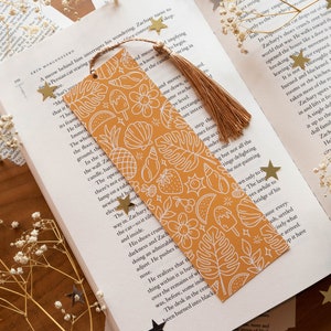 Könnte beinhalten: Ein gelbes Lesezeichen mit einem weißen Design aus tropischen Früchten, Blumen und Blättern. Das Lesezeichen hat einen Quasten an der Oberseite.