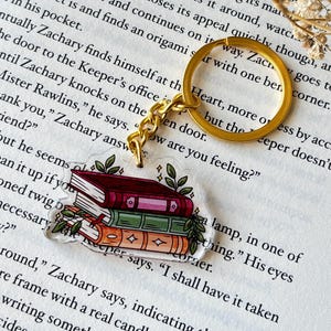 May include: A clear acrylic keychain featuring a stack of three books in shades of red, green, and orange, adorned with green leaves. The keychain has a gold-colored ring and chain.