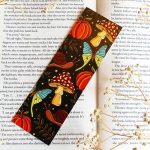Puede incluir: Marcapáginas decorativo con fondo negro y coloridas ilustraciones de calabazas, setas, caracoles y mariposas. El marcapáginas rectangular está sobre un libro abierto.