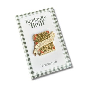 Può includere: Una spilla in smalto a forma di libro con copertina verde, motivo a foglie dorate, angoli rossi e un nastro color crema al centro con la scritta ALWAYS READING. La spilla è fissata su un cartoncino bianco con un bordo a quadretti vichy verdi.