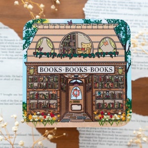 May include: A whimsical illustration of a bookstore with a sign that reads "BOOKS BOOKS BOOKS". The storefront has two windows filled with books and a door with a wreath. There are flowers and greenery around the building.