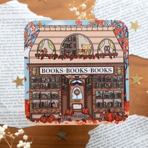 Op de afbeelding: Een eigenzinnige illustratie van een boekwinkel met een bruine bakstenen gevel en een bord met de tekst "BOOKS BOOKS BOOKS". De winkel heeft grote ramen vol boeken, een deur met een krans en een pompoen op de stoep. Het gebouw is omringd door herfstbladeren en een blauwe lucht.