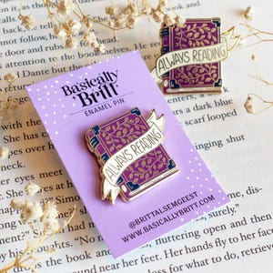 May include: Purple enamel pin shaped like a book with gold trim and a banner that reads "ALWAYS READING." The pin is on a lavender card with the text "Basically Britt ENAMEL PIN." A second pin is visible in the background.