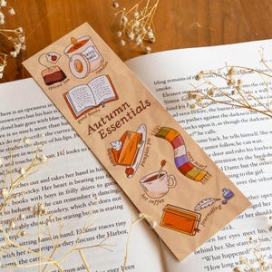 Puede incluir: Un marcapáginas beige con la frase "Autumn Essentials" e ilustraciones de una vela, tarta de calabaza, café y una bufanda. El marcapáginas está sobre un libro abierto.