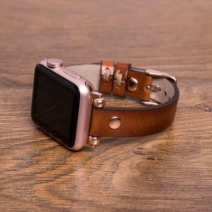 Könnte beinhalten: Eine Smartwatch mit einem roségoldenen Metallgehäuse und schwarzem Display, befestigt an einem braunen Lederarmband. Das Armband verfügt über sichtbare Nähte, eine Metallschnalle und einen Metallnietverschluss, verbunden durch roségoldene Beschläge.