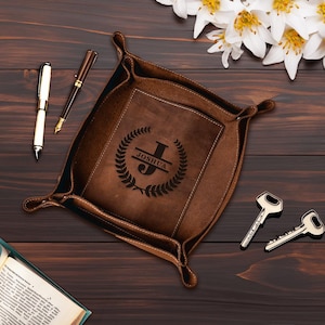 May include: A brown leather catchall tray with a personalized monogram. The monogram features the name "Joshua" and the initial "J" in a laurel wreath design.