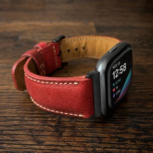 May include: A smartwatch with a matte black casing and a vibrant red leather strap featuring visible cream-colored stitching. The digital display shows the time 12:58, the date Wednesday 28, and health tracking icons including steps, heart rate, and calories.