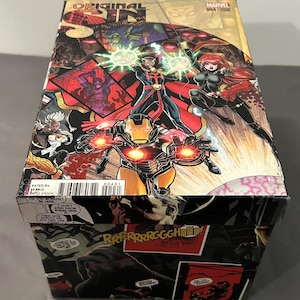 Puede incluir: Una caja de cómic con el título "ORIGINAL SIN" y personajes de Marvel, incluyendo a Iron Man, Doctor Strange y Black Widow. La caja tiene un diseño colorido con escenas de acción y el texto "RATED T".