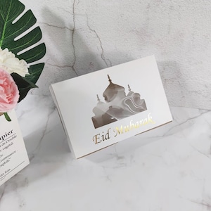 Könnte beinhalten: Weiße Geschenkbox mit einem Ausschnitt in Form einer Moschee und dem Text "Eid Mubarak" in goldenen Lettern.