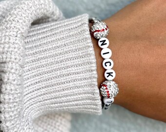 May include: A white and red baseball themed bracelet with the name "NICK" spelled out in black letters.