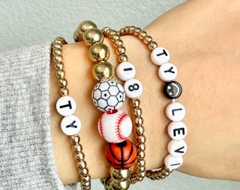May include: Four gold beaded bracelets with white letter beads spelling "TY" and "LLEVI" and a smiley face bead. The bracelets also feature a white soccer ball bead, a white baseball bead, and a basketball bead.