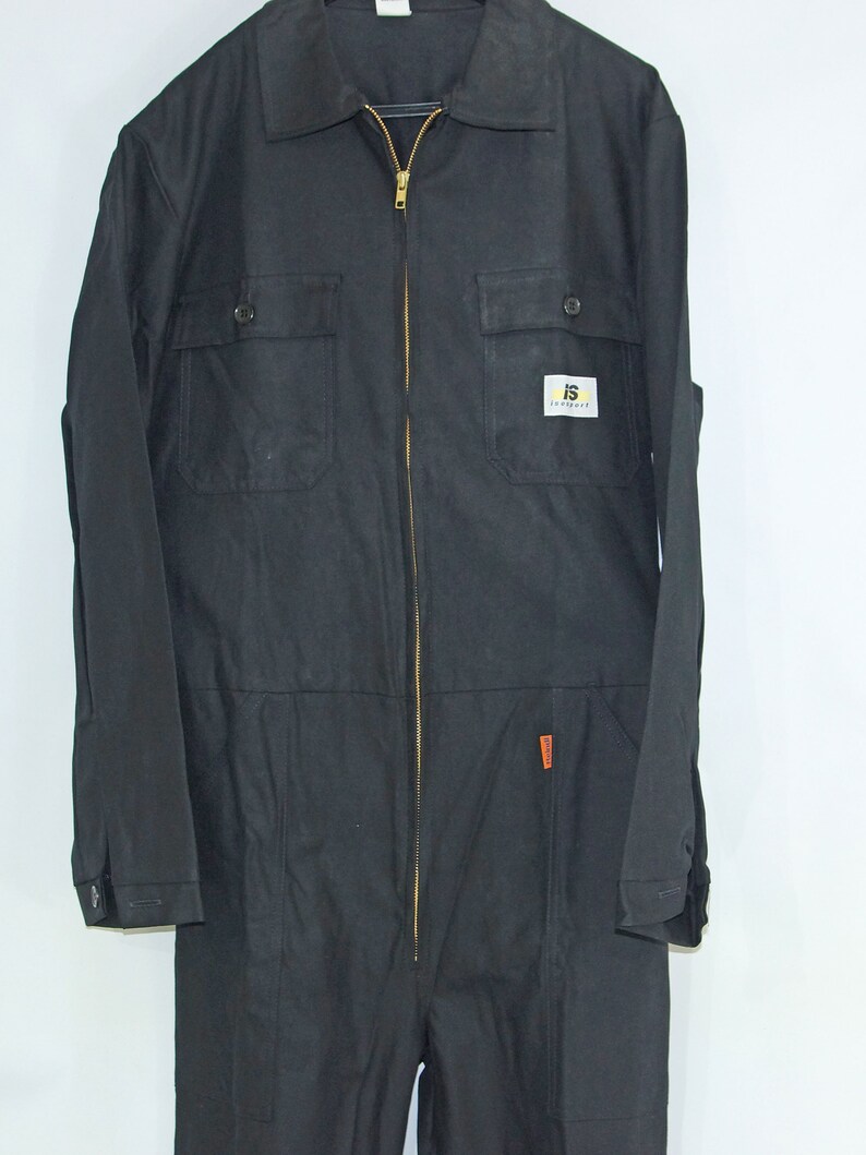 May include: Black workwear jumpsuit with a zipper closure and two chest pockets. The jumpsuit has a small yellow and black logo patch on the chest that says "IS Workwear".