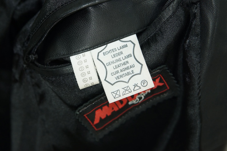 May include: A black leather jacket with a white label that reads "Echtes Lamm Leder, Genuine Lamb Leather, Cuir Agneau Veritable". The label also has a care symbol and a "P" symbol. The jacket has a zippered pocket and a red label that reads "Mad" with a stylized bird logo.