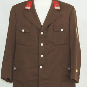 May include: A brown military uniform jacket with silver buttons and red and white collar patches. The jacket has two flap pockets on the front and a patch on the right sleeve.