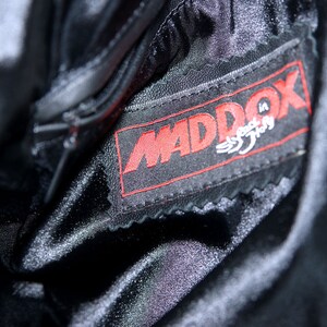 May include: Close-up of a black leather jacket with a red "MADDOX" label sewn on the inside. The label has the words "Speed in Style" written in white.