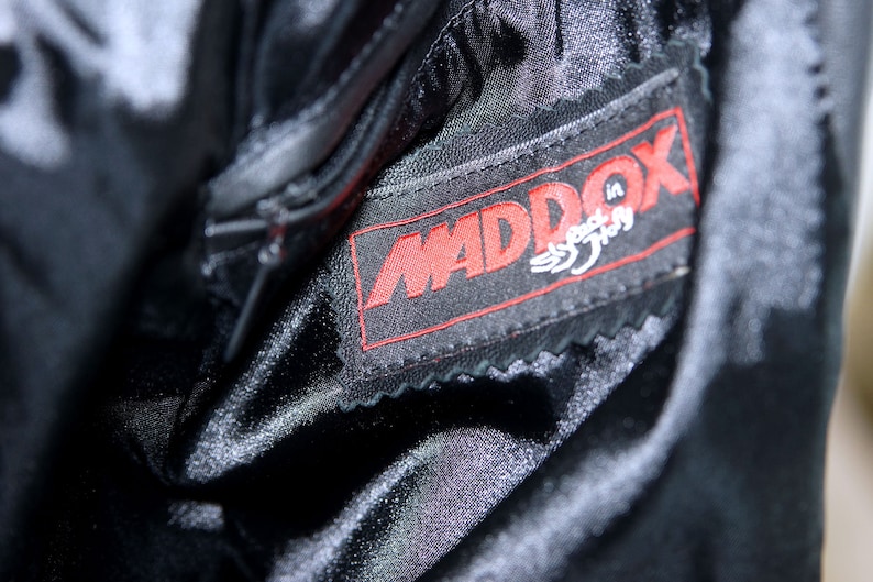 May include: Close-up of a black leather jacket with a red "MADDOX" label sewn on the inside. The label has the words "Speed in Style" written in white.
