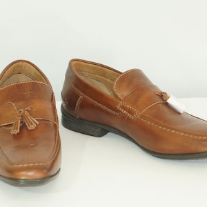 May include: A pair of brown leather loafers with tassel detailing. The shoes feature a classic design with visible stitching and a dark sole. The loafers are displayed on a white surface, highlighting their polished appearance.